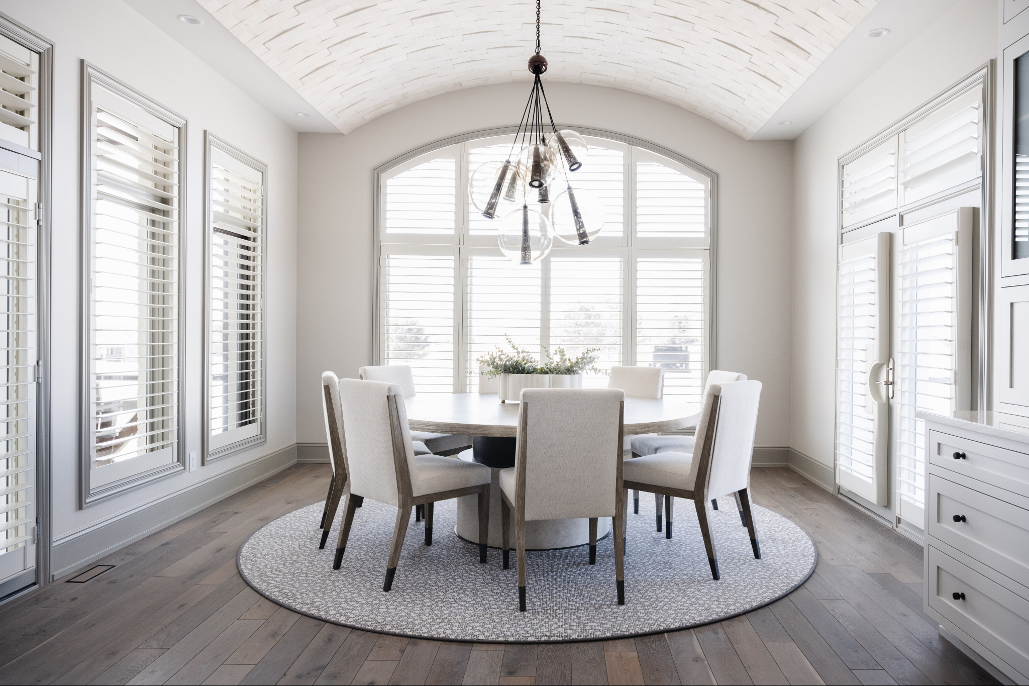 Bright, modern dining room designed by interior designers Omaha NE, featuring a round table with upholstered chairs, vaulted white ceiling, statement chandelier, and large windows with plantation shutters creating a clean, elegant interior design aesthetic.